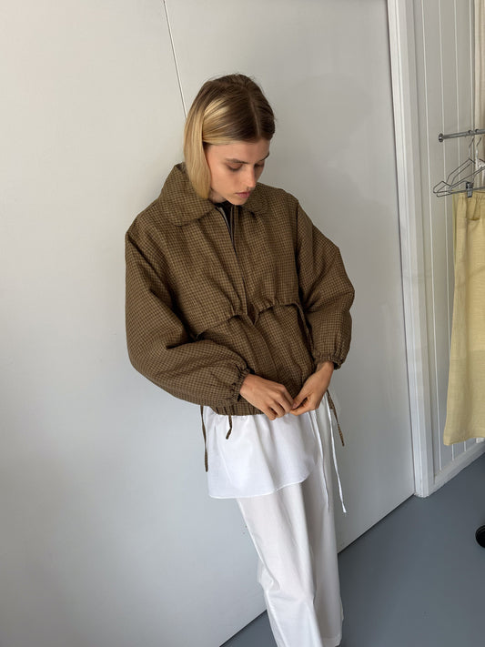 A woman with blonde hair adjusts the drawstrings on the Double Layered Jacket - Pine Check, featuring recycled polyester filling, worn over a long white shirt and white pants. She stands indoors by a white wall and door, next to a clothes rack.