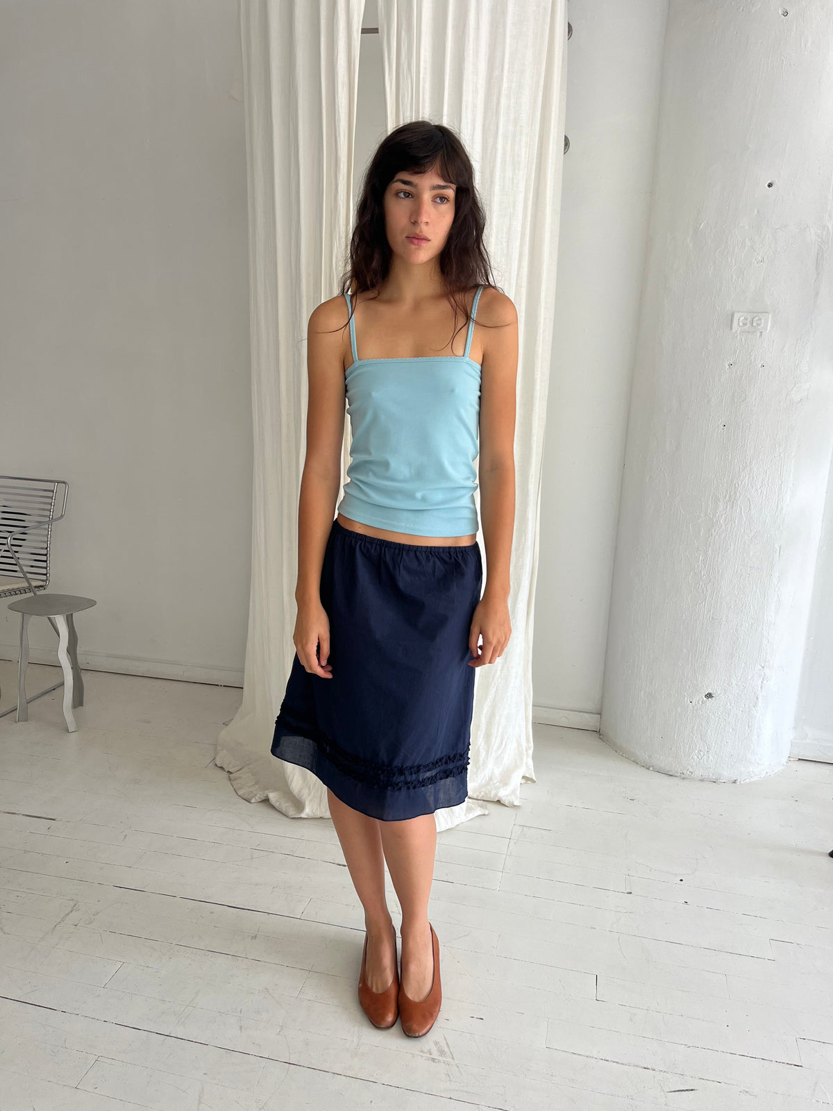 A woman stands against a white curtain, wearing the Daytime Skirt - Navy by Deiji Studios, a cotton voile midi skirt with a straight silhouette and double frill detailing at the hem, styled with the Scallop Tank in Sky.