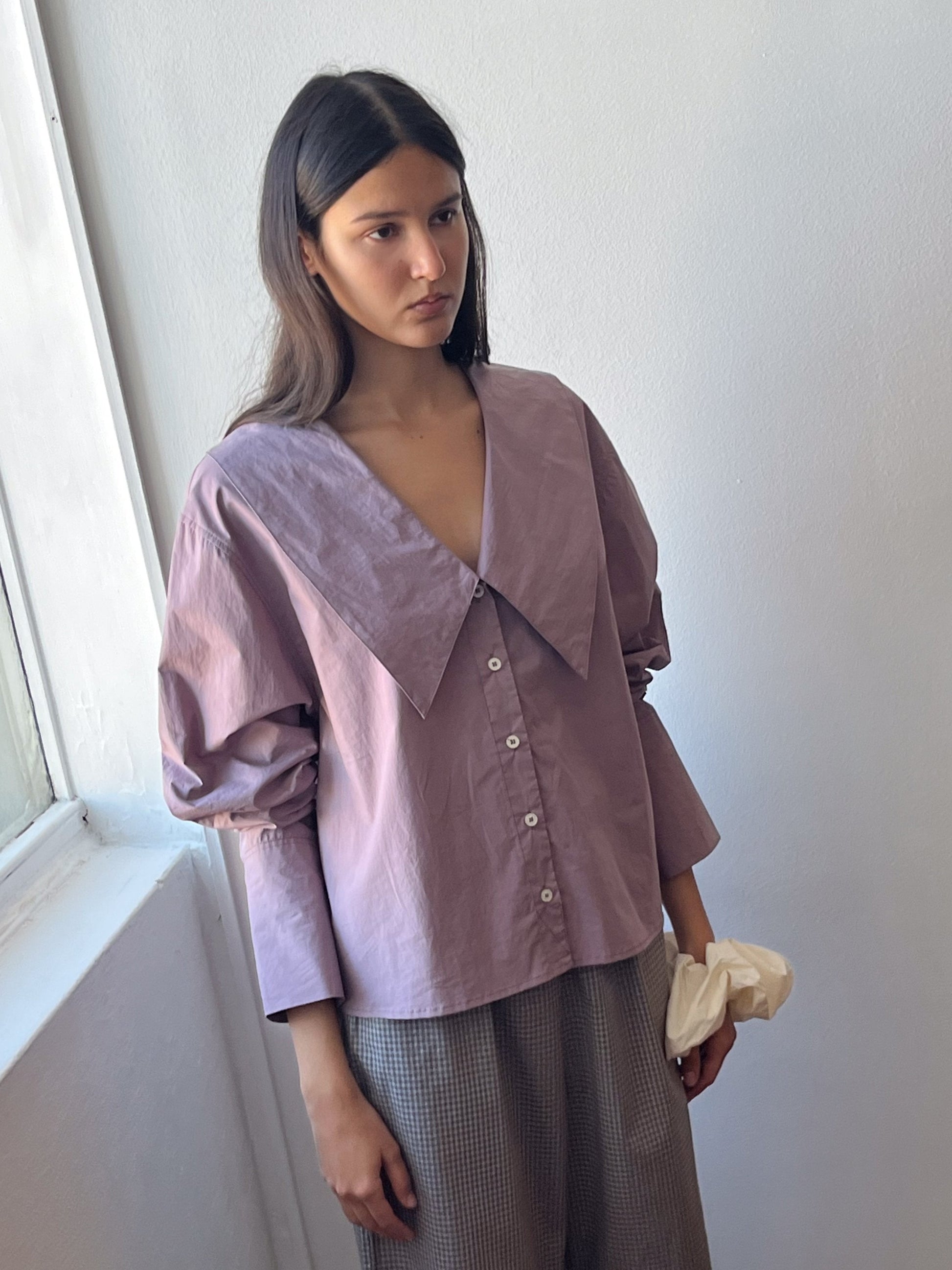 A woman with long brown hair stands by a sunlit window, wearing the Oversized Collared Shirt in mauve with puffed sleeves and a large collar, styled with gray checkered pants. She holds a white cloth and appears thoughtful.