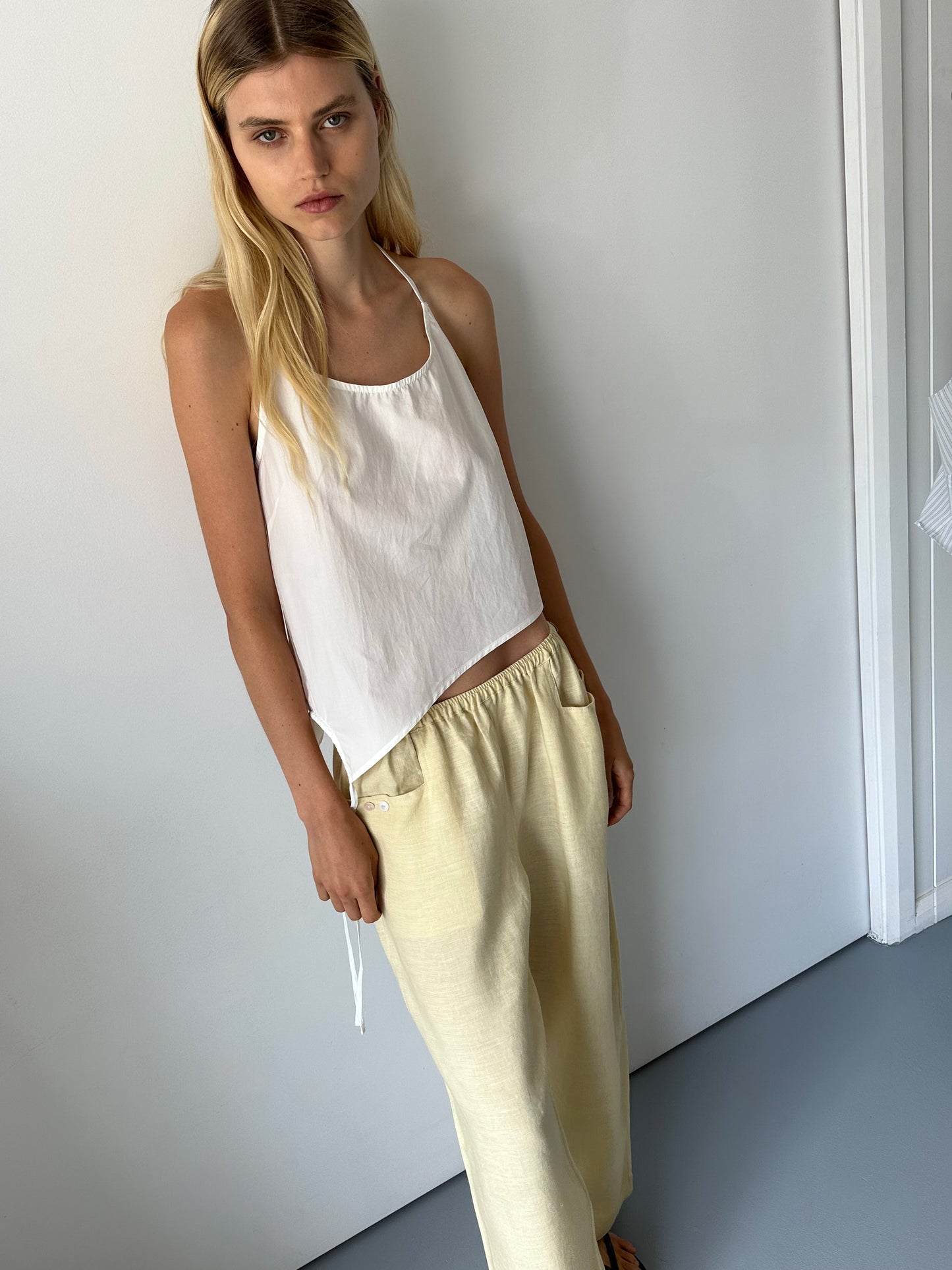 A woman with long blonde hair wearing a white sleeveless top and the Linen Square Pocket Pant in Butter Yellow stands indoors against a plain white wall, looking at the camera with a neutral expression.