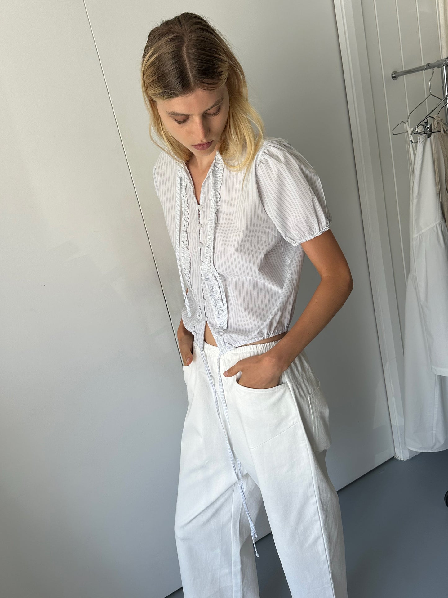 A person with blonde hair wears the Double Frill Top - Sage Stripe by 0, tucked into high-waisted white pants. Indoors, they look down with hands in pockets near white walls and hanging clothes.