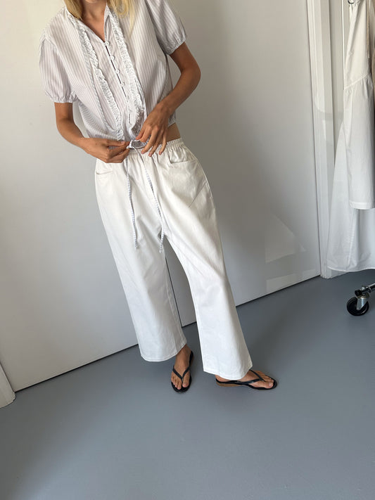 A person stands indoors on a gray floor, adjusting the drawstring ties of a short-sleeved, light-striped blouse. Theyre wearing 0s Square Pocket Pant in white with black flip-flops. Their face is out of frame.