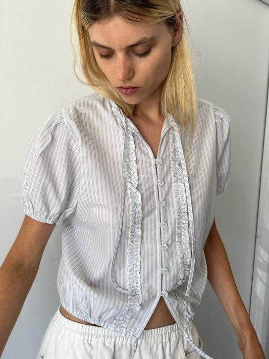 A woman with blonde hair wears the Double Frill Top - Sage Stripe by 0, featuring short sleeves and subtle pinstripes. She looks down with a neutral expression against a plain background.