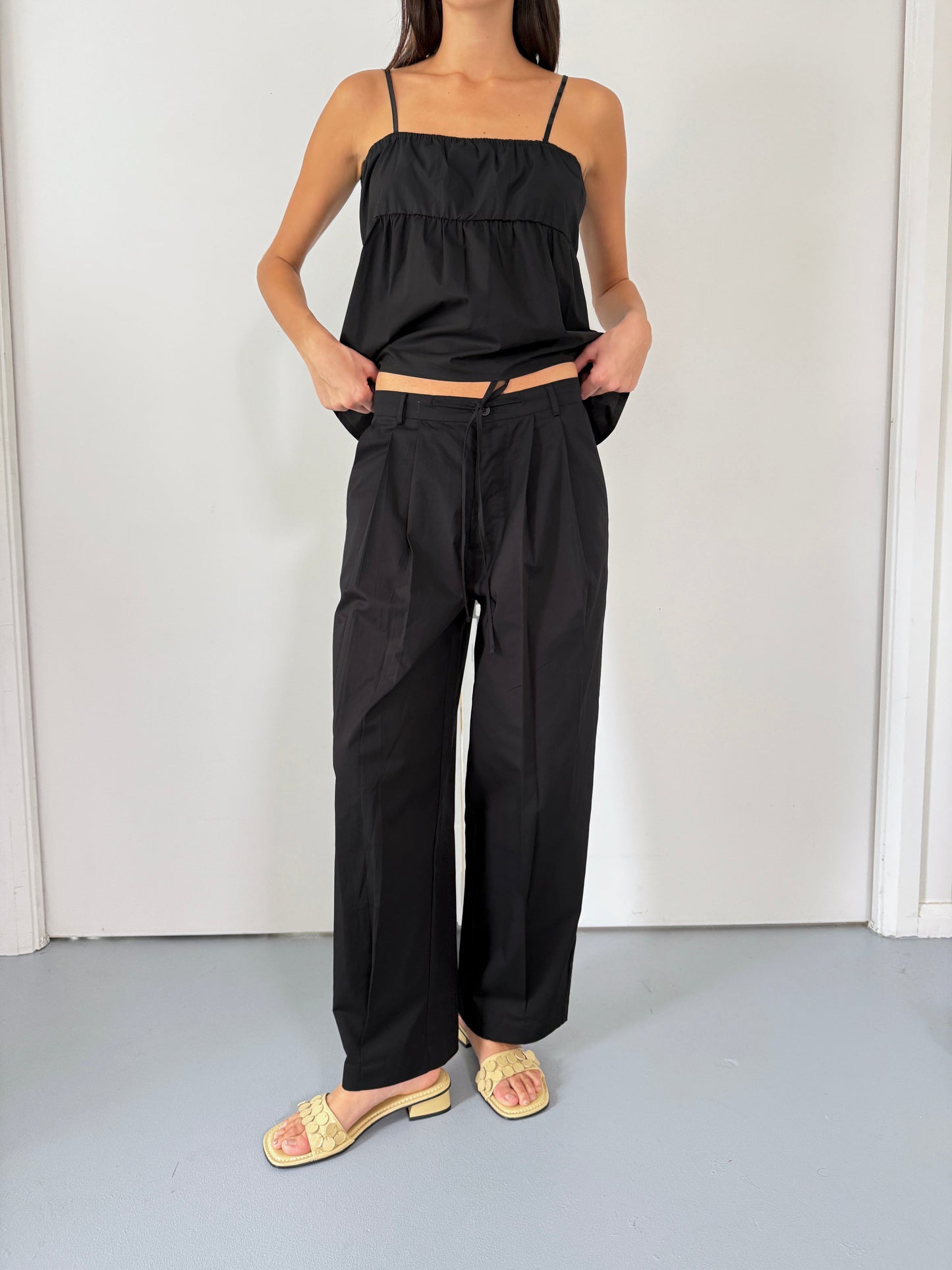 female model wears the turn pant in black, a relaxed tailored pant features soft pleating at the front, buttoned waistband and adjustable inner drawcord. designed with hidden side seam pockets and pockets at the back, made from 100% GOTS certified organic cotton paired with a black top