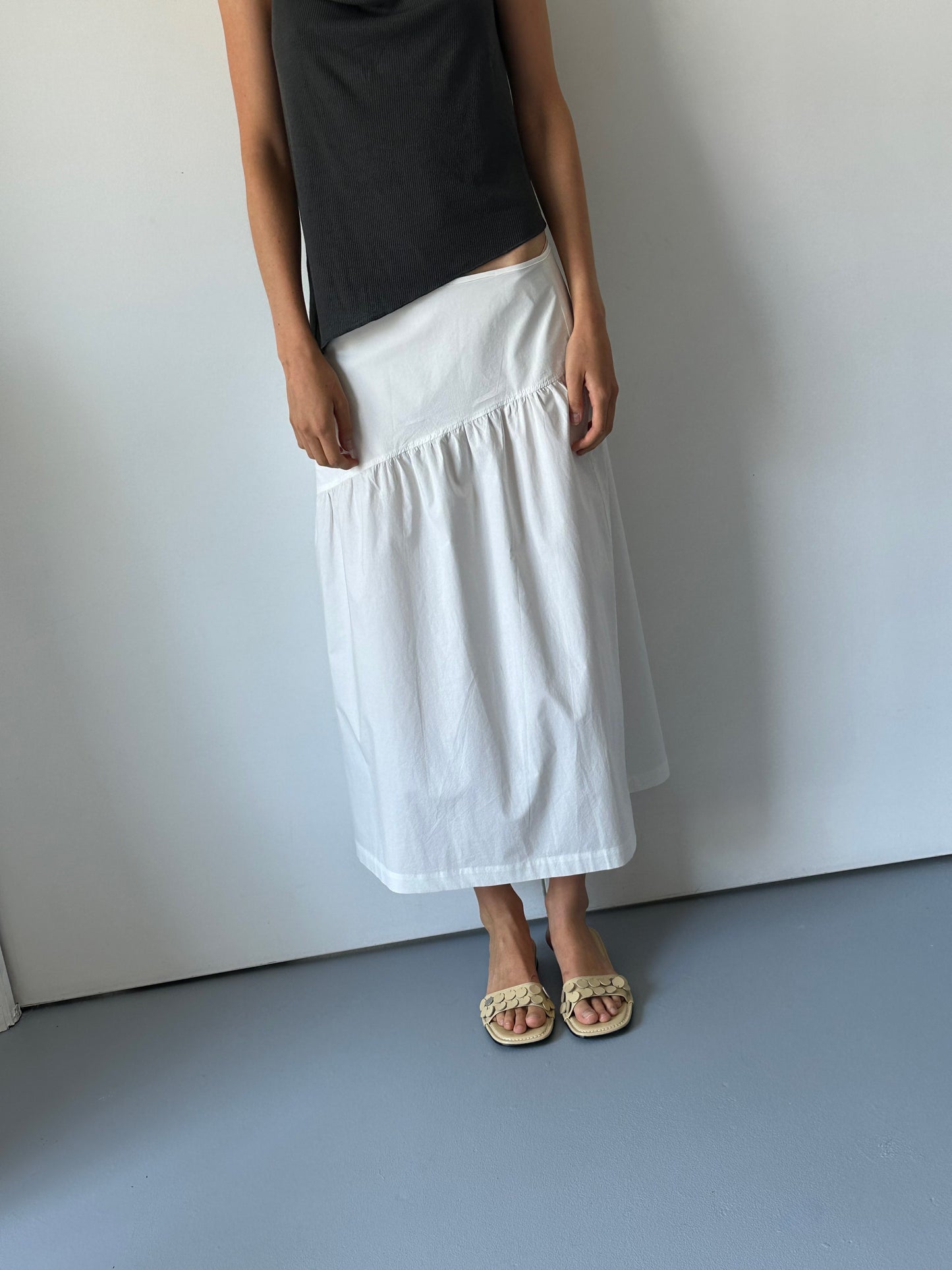 A person wearing a sleeveless black top, the Angle Skirt - White in organic cotton, and beige braided sandals stands against a plain light-colored wall and gray floor. Their face is not visible.