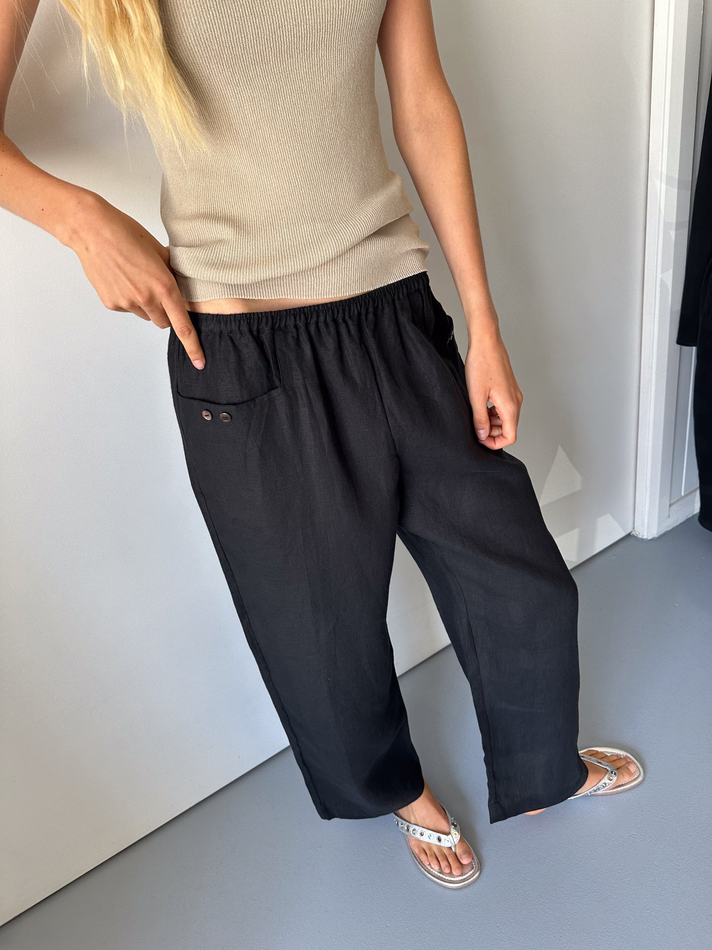 A person in a beige sleeveless top, flip-flops, and 0s Linen Square Pocket Pant in Black with buttons by the pocket stands indoors, pointing to the pants waistband.