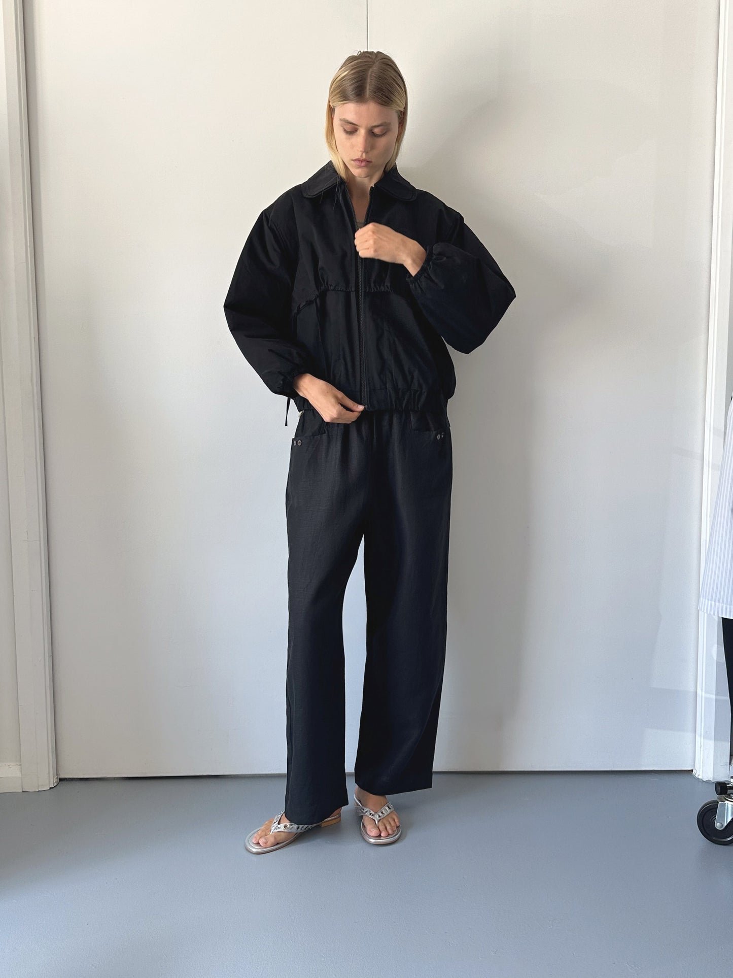 A person stands indoors against a white wall, wearing a loose black jacket and 0 Linen Square Pocket Pant - Black with silver sandals. Their head is slightly tilted down, and their right hand adjusts their jacket.