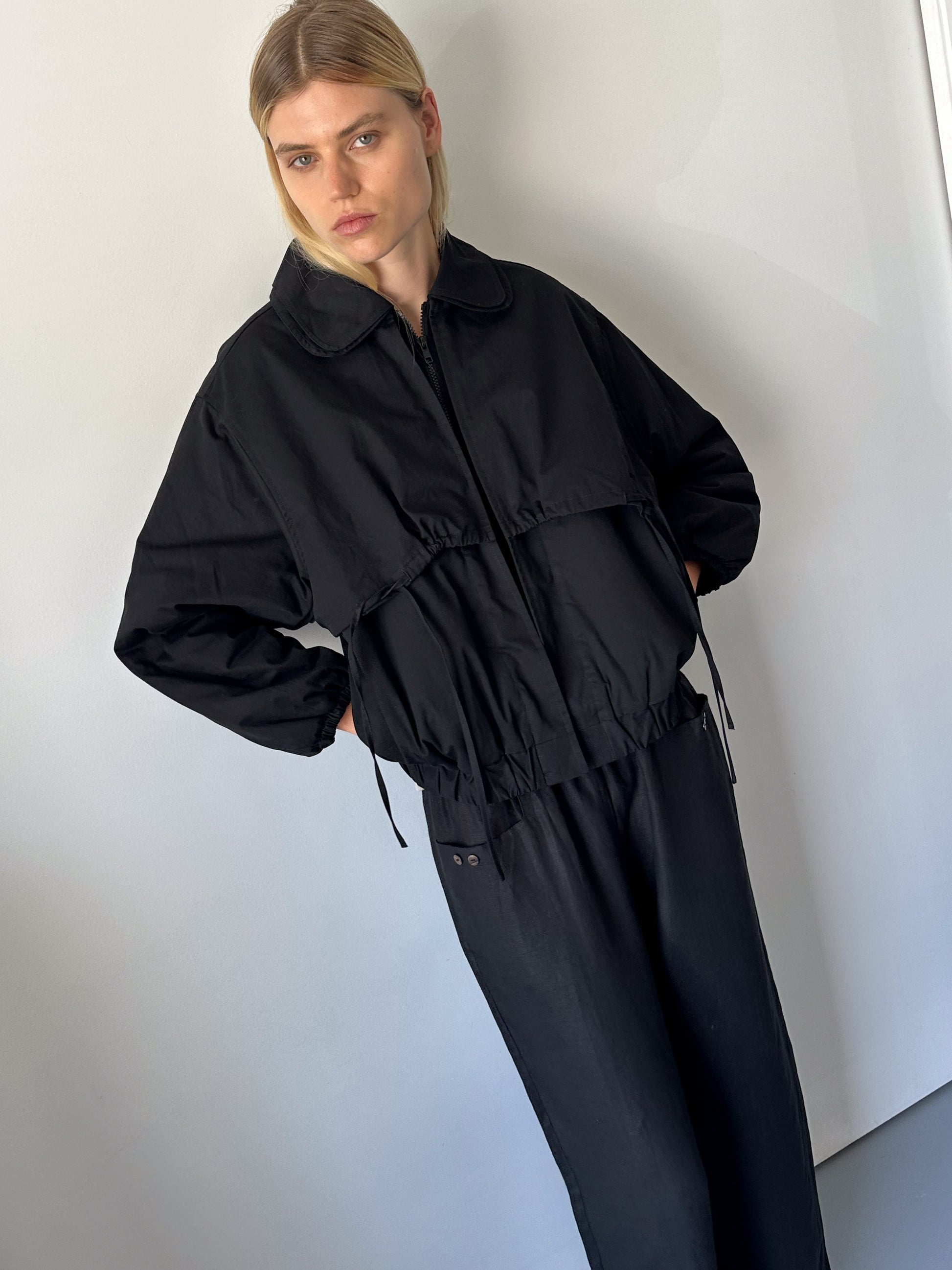 A person with straight blond hair stands against a light gray wall, wearing the 0 Double Layered Jacket - Black and matching black pants, looking slightly to the side with a neutral expression.
