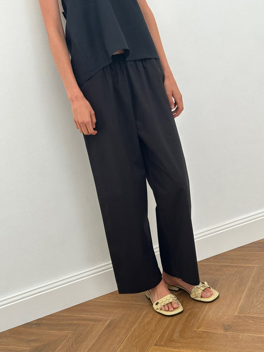 A person stands indoors on wooden flooring, wearing a sleeveless black top, the Ease Trouser - Black from Deiji Studios, and light yellow slide sandals with a bow detail. Their face is not visible.
