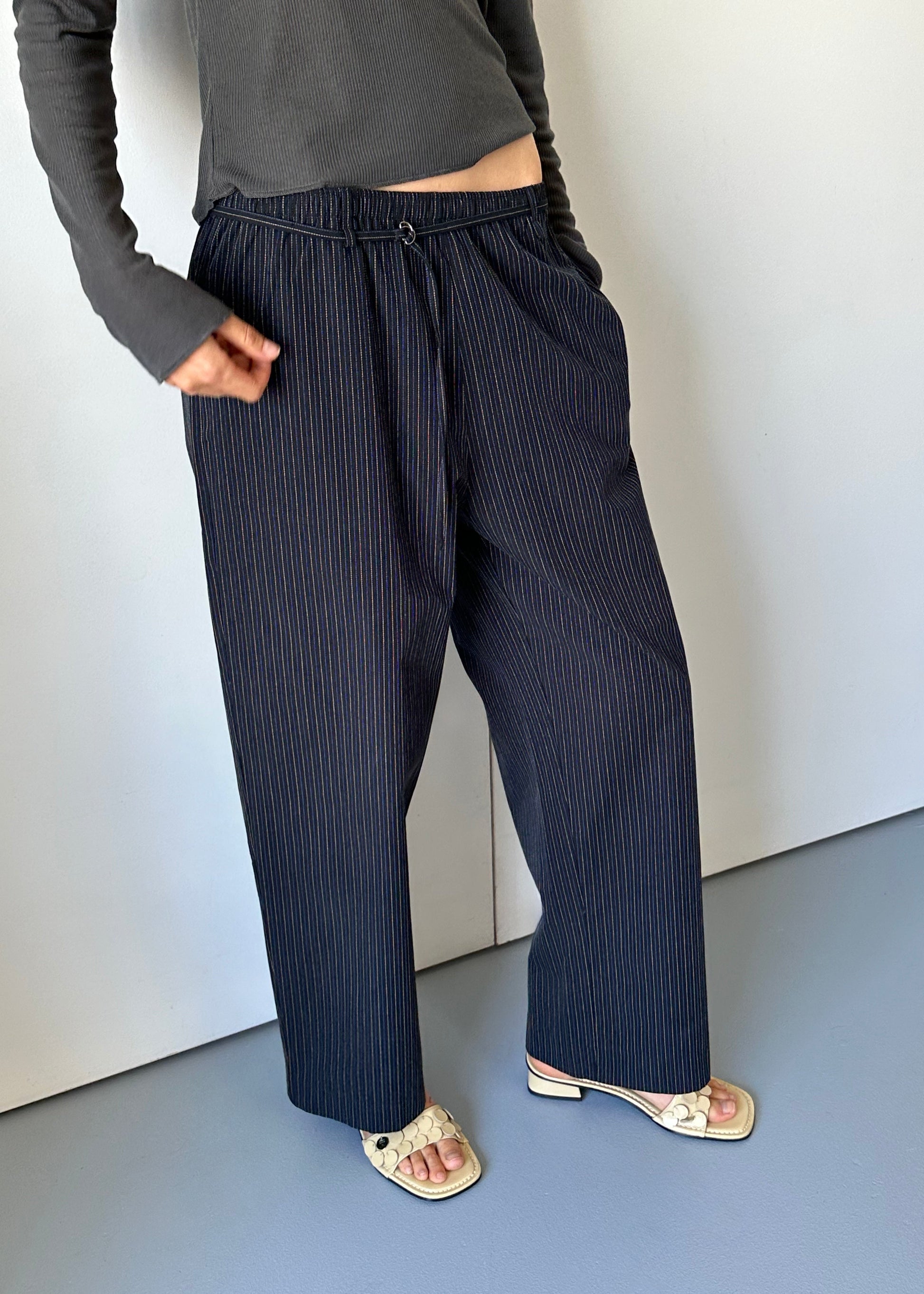 Someone stands against a light wall with hands in pockets, wearing the 0 Loop Pant - Ink Butter Stripe, a relaxed pinstripe trouser, paired with a dark gray long-sleeve top and cream heeled sandals. The head is not visible.