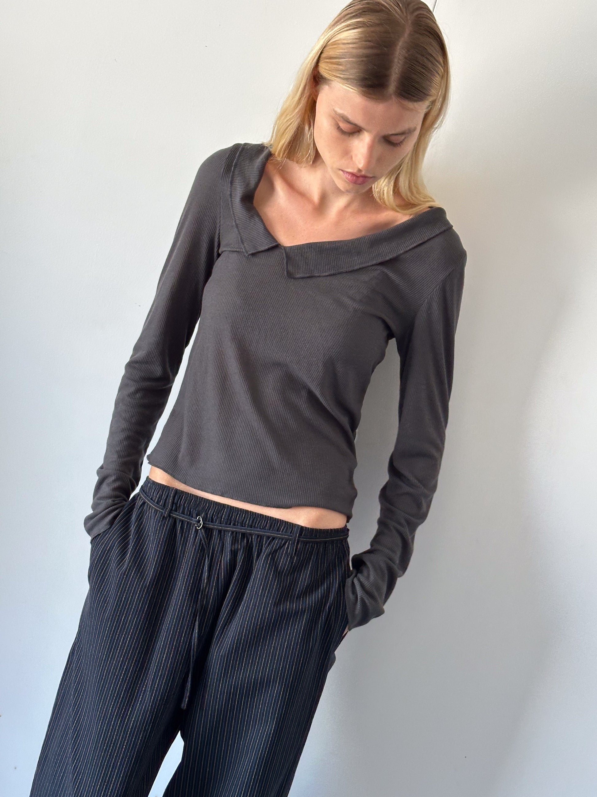 A person with straight blonde hair looks down, wearing the Cross Collar Top - Night Blue with an asymmetrical neckline and loose, pinstriped black pants, standing against a plain light-colored wall.