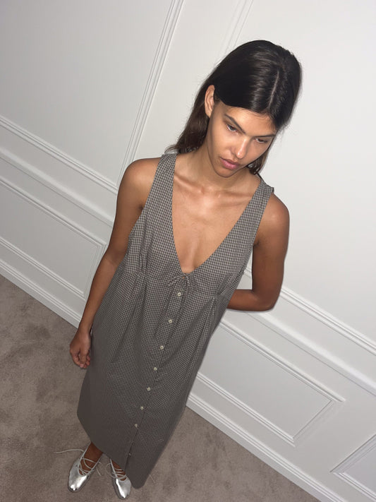 A woman with long dark hair wears the Tuck Tie Dress—a sleeveless, button-down, V-neck style with a small check pattern—and silver ballet flats, standing indoors on a carpeted floor against a white paneled wall.