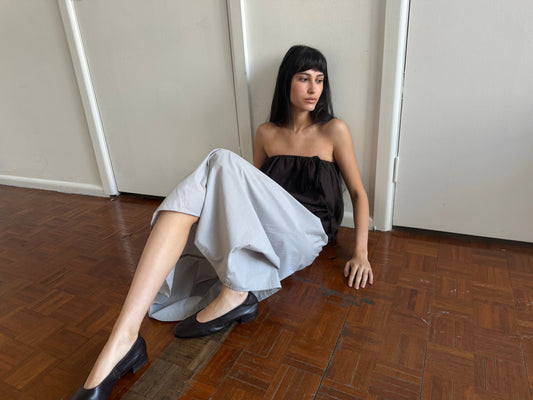 A woman with dark hair and a fringe sits on the floor against a door wearing the Bubble Top - Brown Black Stripe by Deiji Studios, a strapless dark brown bubble-hem top, with a pale grey skirt and black flats.
