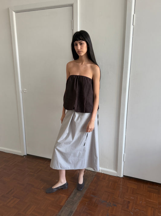 A woman with dark hair and a fringe stands in a room wearing the Wrap Skirt - Cloud Stripe by Deiji Studios, a pale grey-blue fine-striped wrap midi skirt, with a brown strapless bubble top and grey flats.