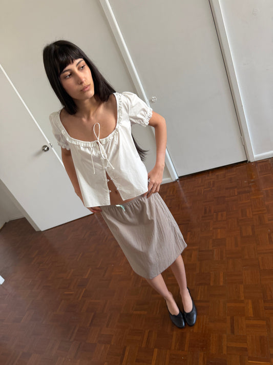 A woman with dark hair and a fringe stands in a room wearing an open white frill top and the Basic Skirt - Grey by Deiji Studios, a grey cotton voile midi skirt, with black flats.