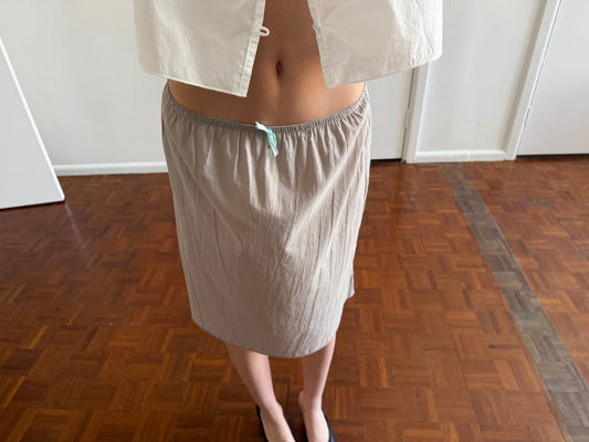 A cropped view of the Basic Skirt - Grey by Deiji Studios, a grey cotton voile midi skirt with an elasticated drawstring waist, worn with a white top and black flats in a room.
