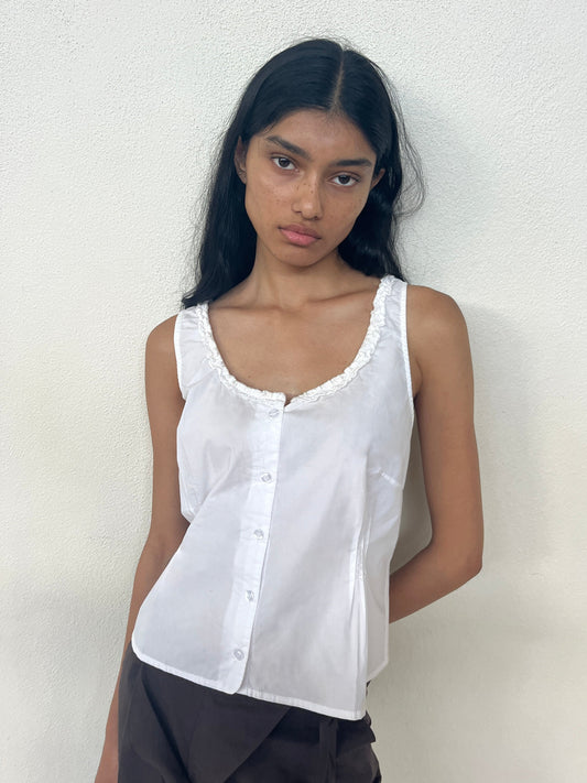 A model with long dark hair wears the Ruffle top, a white sleeveless button-front style with a ruffled scoop neckline, paired with dark trousers and standing against a white textured wall.
