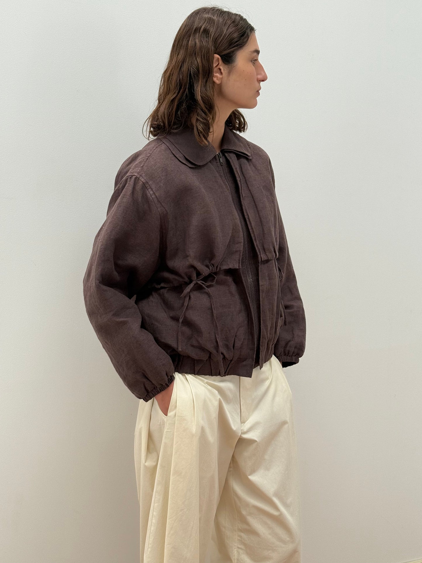 A person with wavy brown hair stands sideways against a plain wall, wearing the Double Layered Jacket - Bark by Deiji Studios with a drawstring waist and off-white wide-leg pants, hands in pockets.