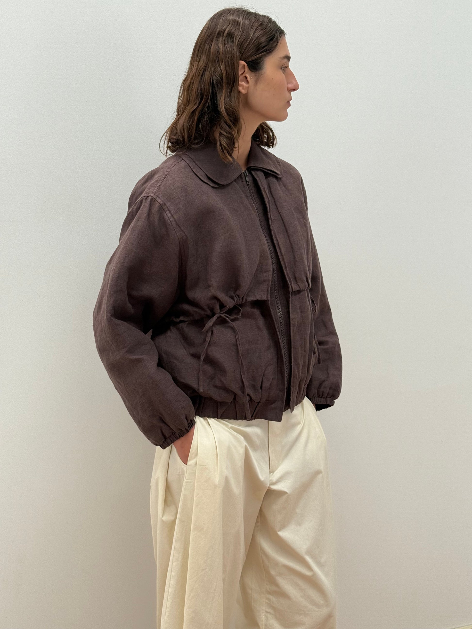 A person with wavy brown hair stands sideways against a plain wall, wearing the Double Layered Jacket - Bark by Deiji Studios with a drawstring waist and off-white wide-leg pants, hands in pockets.