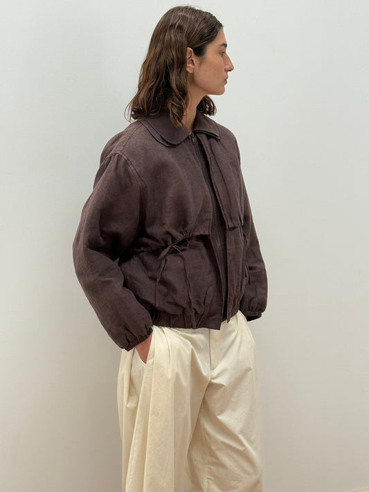 A person with wavy brown hair stands sideways against a plain wall, wearing the Double Layered Jacket - Bark by Deiji Studios with a drawstring waist and off-white wide-leg pants, hands in pockets.