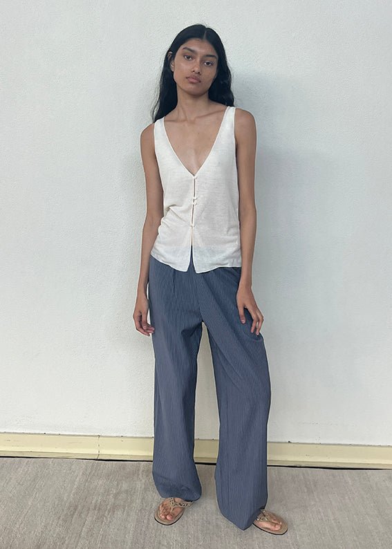 A woman with long dark hair wears the Button Up Knit Tank in Off White—a sleeveless V-neck top—and loose blue pinstriped pants with sandals. She stands against a plain, light-colored wall on a wooden floor, looking slightly toward the camera.
