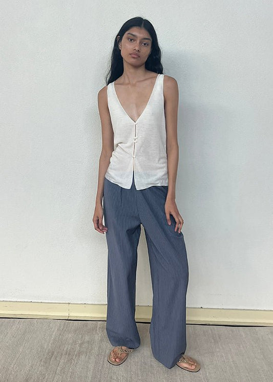 A woman with long dark hair wears the Button Up Knit Tank in Off White—a sleeveless V-neck top—and loose blue pinstriped pants with sandals. She stands against a plain, light-colored wall on a wooden floor, looking slightly toward the camera.