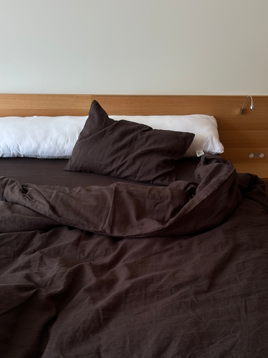 A neatly made bed features dark brown Deiji Studios bedding and a Pillow slip set - Bark, with a white bolster against a wooden headboard. A small reading light is mounted on the wall above the bed.
