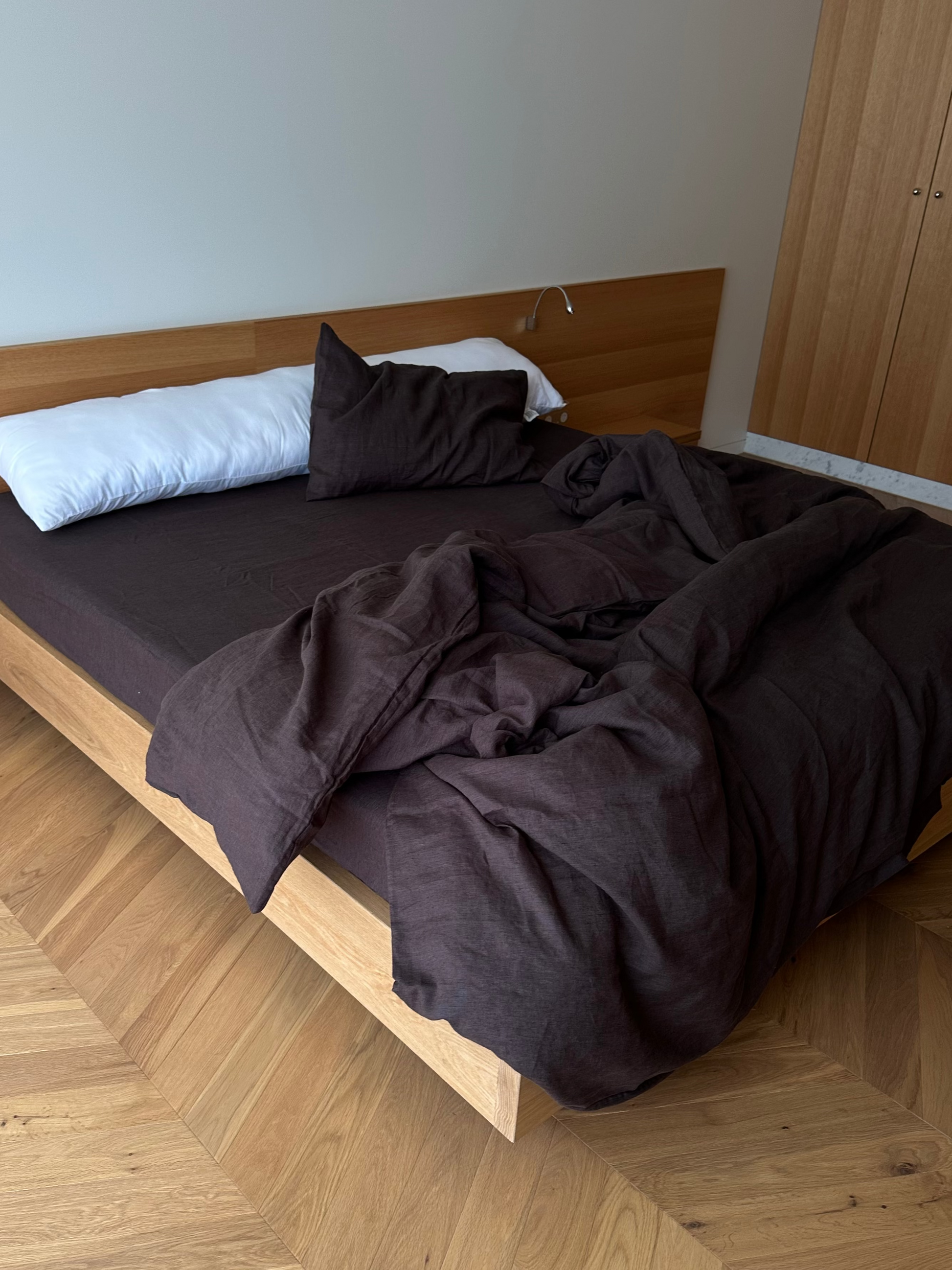 A Duvet cover - Bark US in dark brown covers the bed, paired with a black pillow, matching brown mattress, breathable linen sheets, and a white pillow at the headboard near a built-in wooden wardrobe. The bedding is rumpled and partly hanging off the bed.