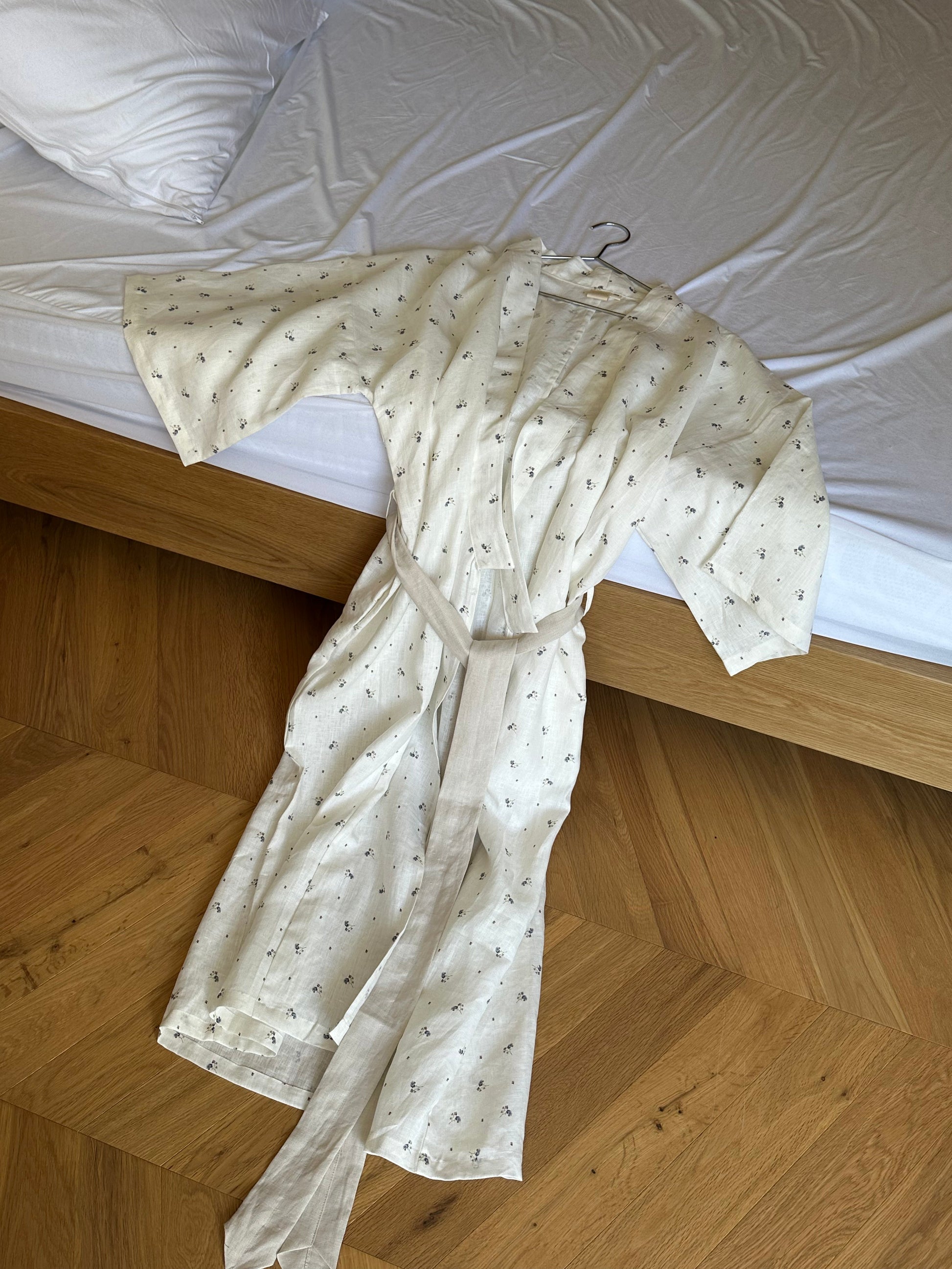 The 02 Robe, a white patterned belted robe with a collar hanger, is displayed on a wooden floor beside an unmade bed with white sheets.