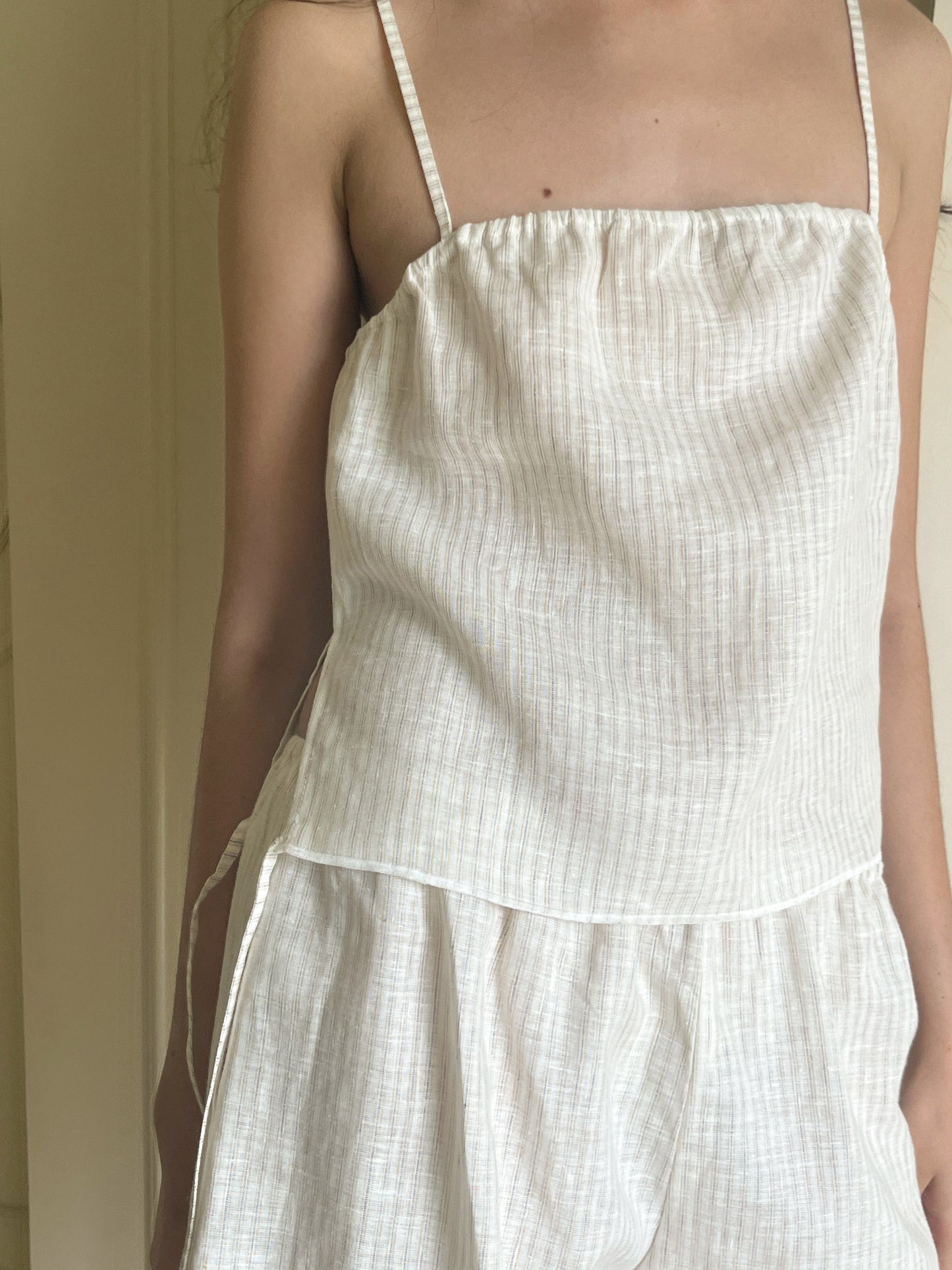 A person wears the Babylock Linen Top - Linen Story Stripe from Deiji Studios, a sleeveless, loose organic linen top with thin straps and subtle stripes. The photo is cropped from shoulders to just above the knees.