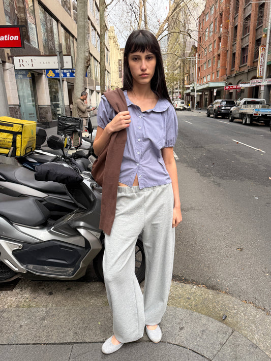 A woman with dark hair and a fringe stands on a city street wearing the Petal Top - Thistle by Deiji Studios, a pale periwinkle petal-collar blouse, with grey marle trousers, a brown shoulder bag and grey flats.