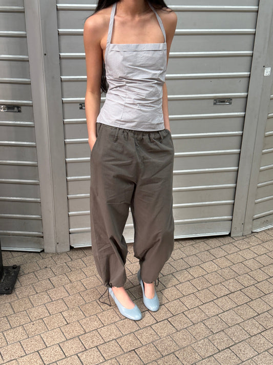 A cropped view of a woman outdoors wearing the Hem Toggle Pant - Moss Stripe by Deiji Studios, moss-green trousers with a toggle-gathered hem, with a pale striped halter top and pale blue heels.