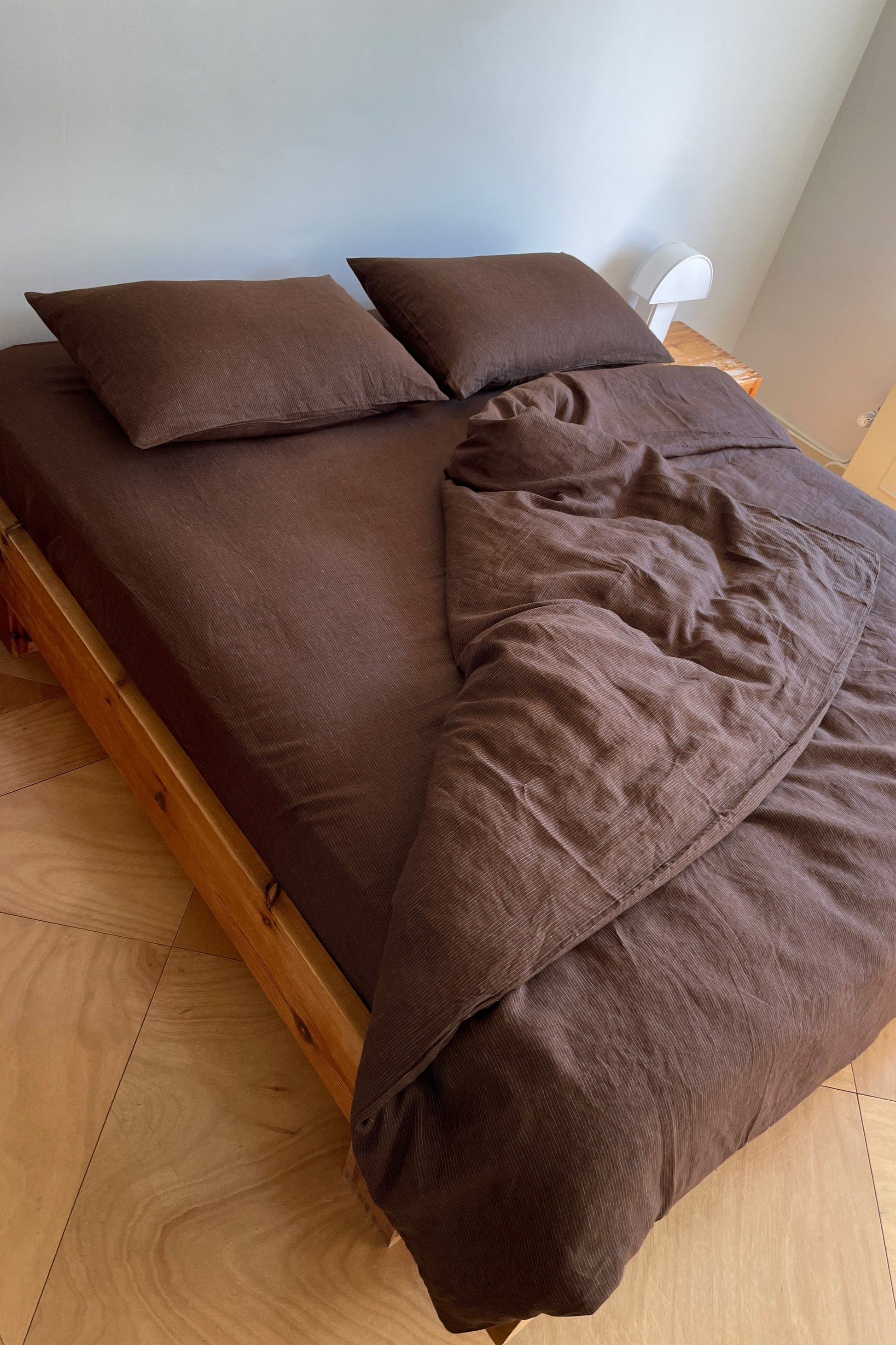 A neatly made wooden bed features Deiji Studios Duvets in dark brown, two pillows, and a matching comforter partially folded back. To the right, a small white lamp sits next to sunlight streaming in from a window.