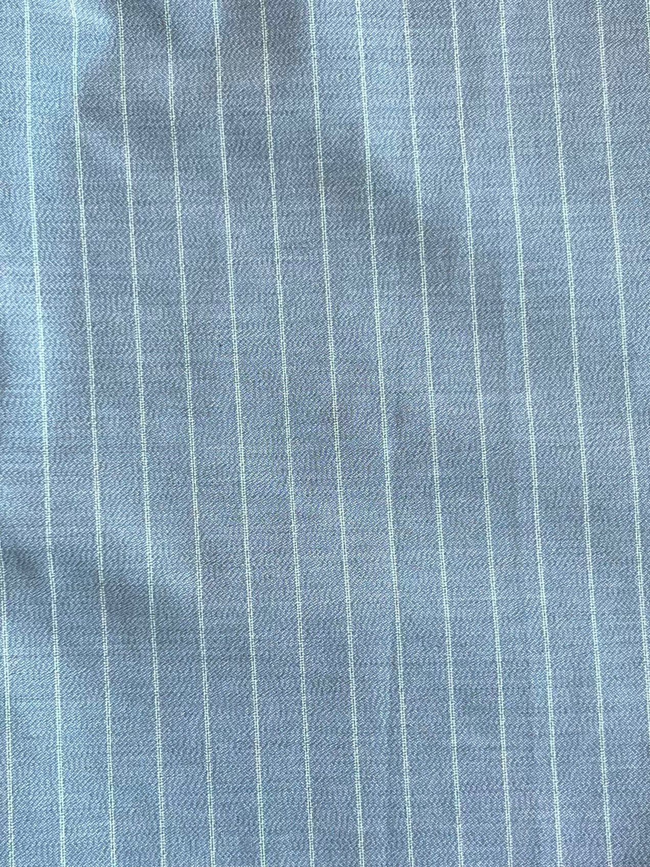 Close-up of light blue Tri Pleat Pure Wool Pant - Steele Stripe by Deiji Studios, featuring RWS wool fabric with thin white vertical pinstripes and subtle wrinkles.