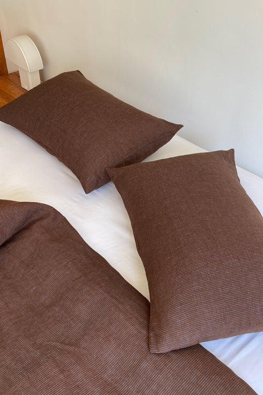 Two brown Pillow Slips and a matching blanket are placed on a white bed. A small white lamp stands on a wooden surface nearby, enhancing the minimalist, modern vibe.