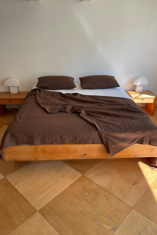 Brown Sheets cover the wooden bed, which has two matching brown pillows and sits against a white wall. Small wooden nightstands with white lamps are on each side. Sunlight shines on the tiled floor.