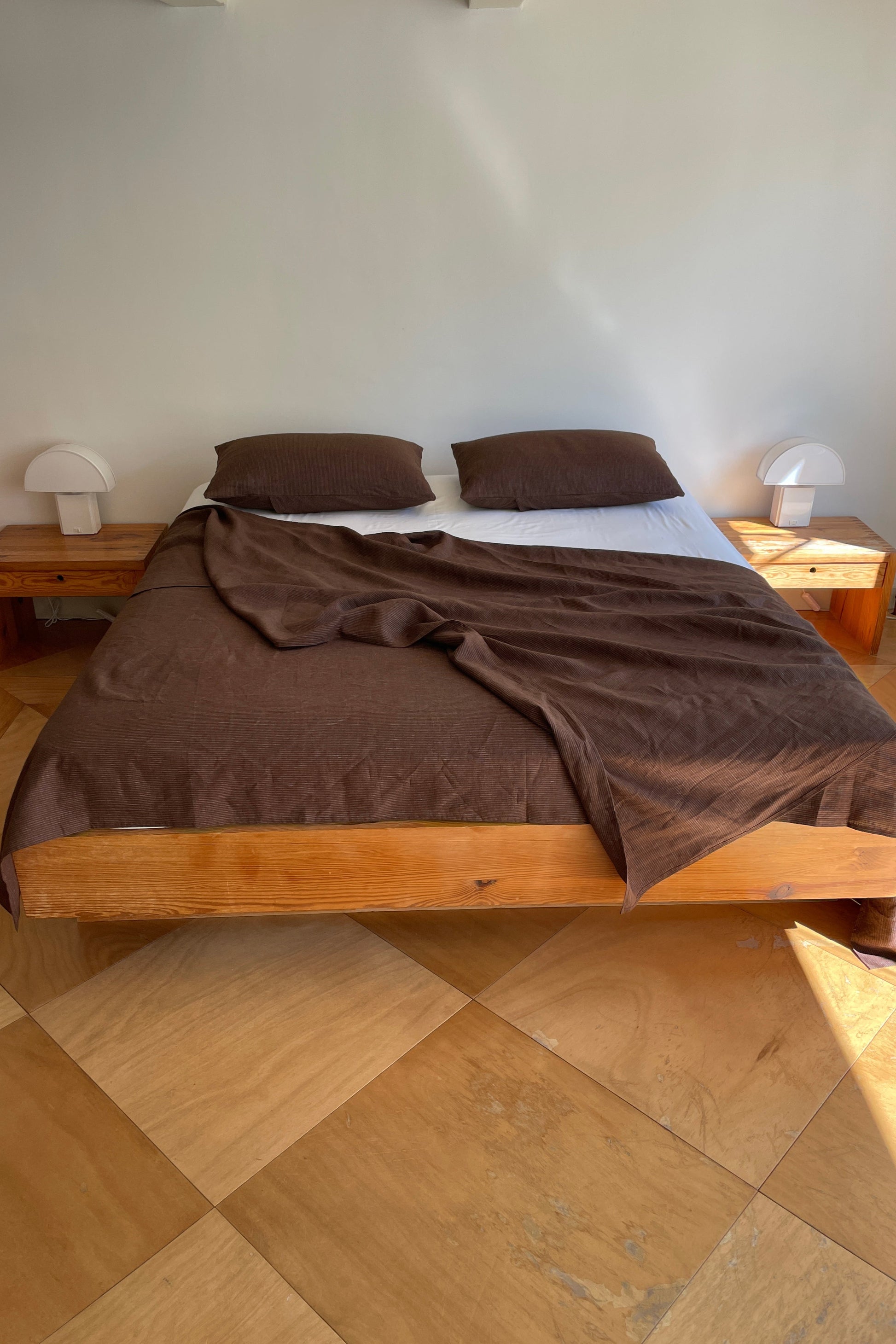 A wooden bed with Sheets - Mahogany Royal Stripe and two pillows stands against a plain white wall. Two small nightstands with white lamps are on either side, and large square tan tiles cover the floor.
