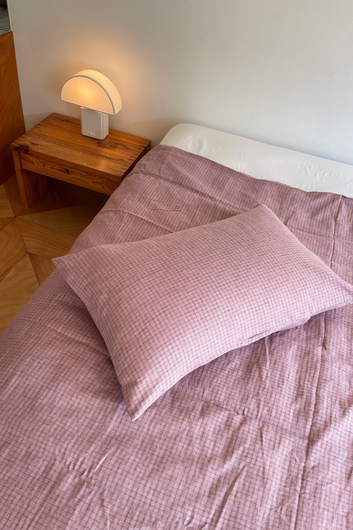 A neatly made bed with light pink checkered Pillow Slips and a matching blanket sits beside a wooden nightstand holding a modern white lamp, all set against a plain white wall.