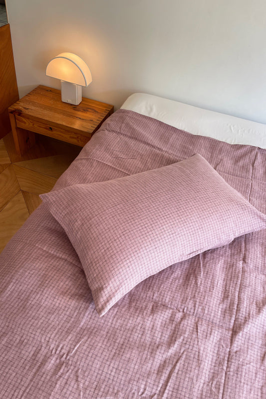 A neatly made bed with light pink checkered Pillow Slips and a matching blanket sits beside a wooden nightstand holding a modern white lamp, all set against a plain white wall.