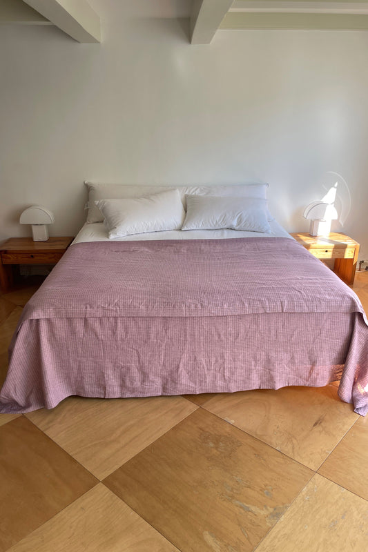 A neatly made bed features white pillows and the Sheets light pink bedspread, centered against a white wall with wooden nightstands and white lamps. The room has light wooden floors and a minimalistic style.