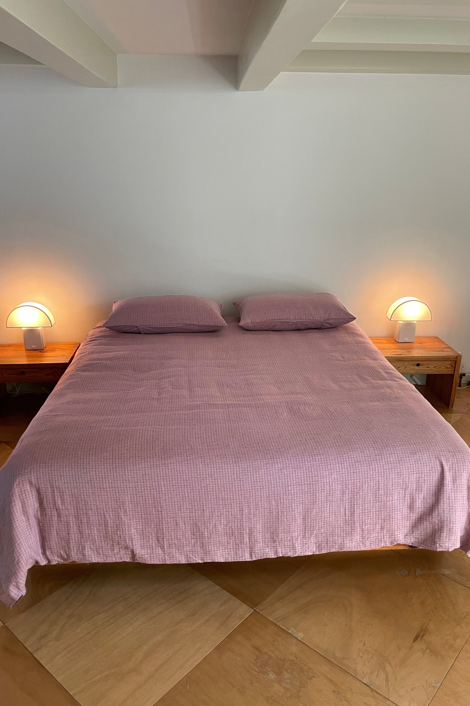 A neatly made bed with a pink Deiji Studios Duvets and two pillows stands against a plain white wall, bordered by wooden nightstands with round white lamps on tan tiled floors.