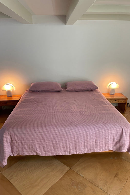 A neatly made bed with a pink Deiji Studios Duvets and two pillows stands against a plain white wall, bordered by wooden nightstands with round white lamps on tan tiled floors.