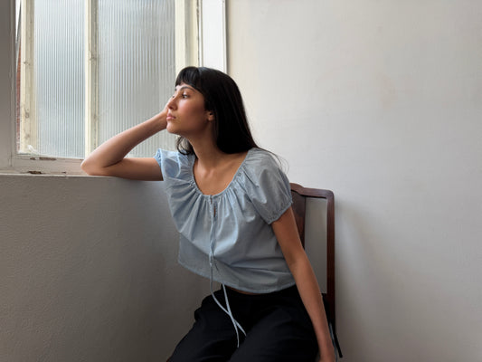 A woman with dark hair and a fringe sits by a window wearing the Tie Top - Blue by Deiji Studios, a pale blue top with a gathered neckline and puff sleeves, with black trousers.