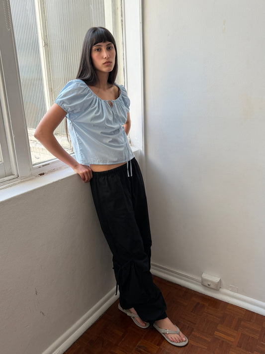 A woman with dark hair and a fringe leans against a windowsill wearing the Tie Top - Blue by Deiji Studios, a pale blue top with a gathered neckline and puff sleeves, with black trousers and flip-flops.