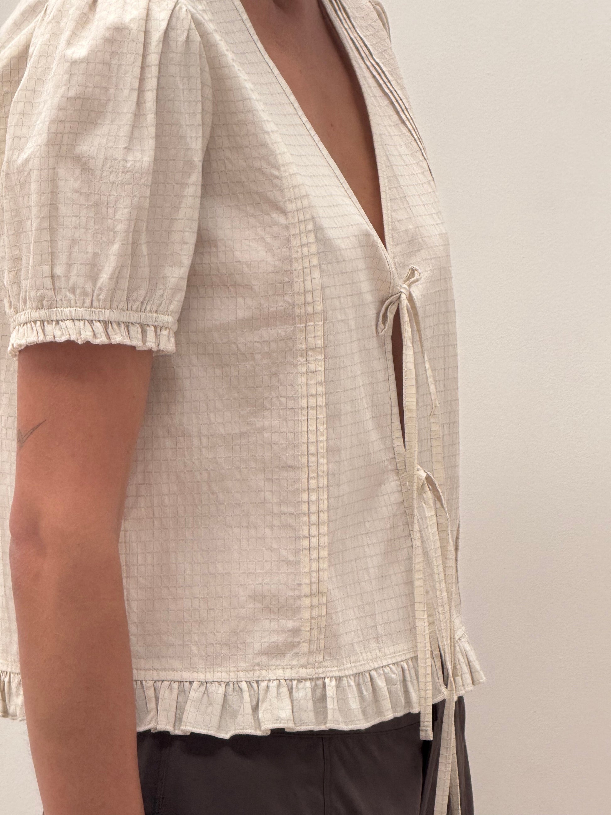 A person wears the Pintuck Top - Bone Check: a light beige, short-sleeved blouse made from organic cotton with a grid pattern, puffed sleeves, ruffled hem, and deep V-neck with tie strings. The background is plain and light-colored.