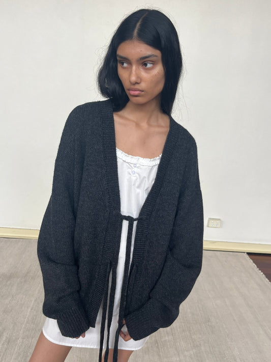 A person with long dark hair stands indoors on a light carpet, wearing the Two Tie Front Cardigan in black over a white dress. They appear thoughtful and gaze slightly to the side against a plain wall.