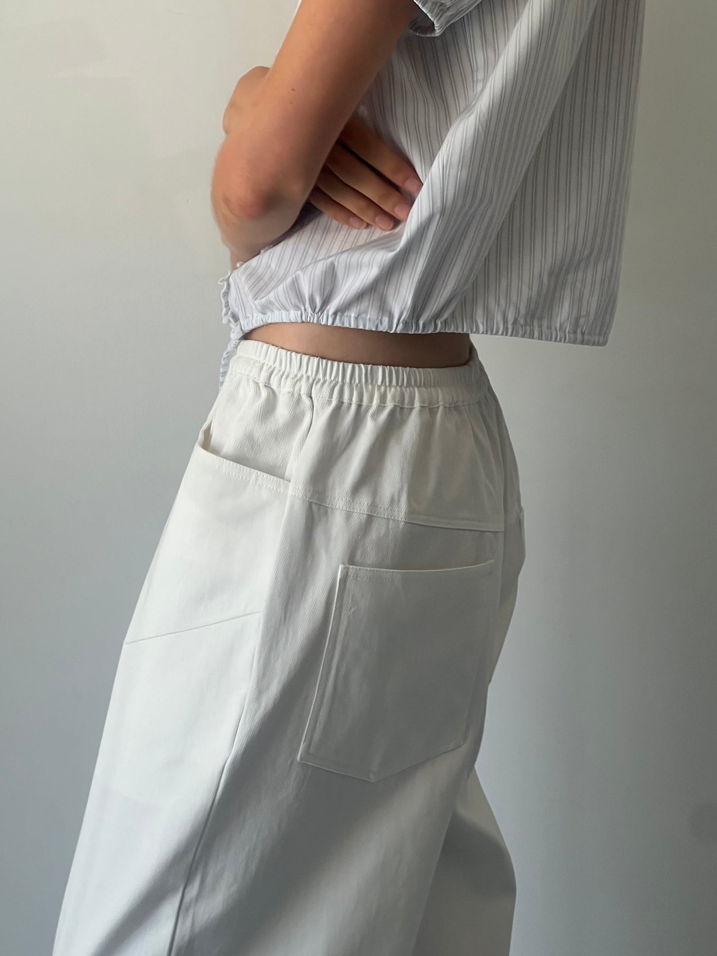 A person stands with arms crossed against a plain background, wearing 0s Square Pocket Pant - White in organic cotton twill, styled high-waisted and wide-leg, paired with a short-sleeve light-striped shirt.
