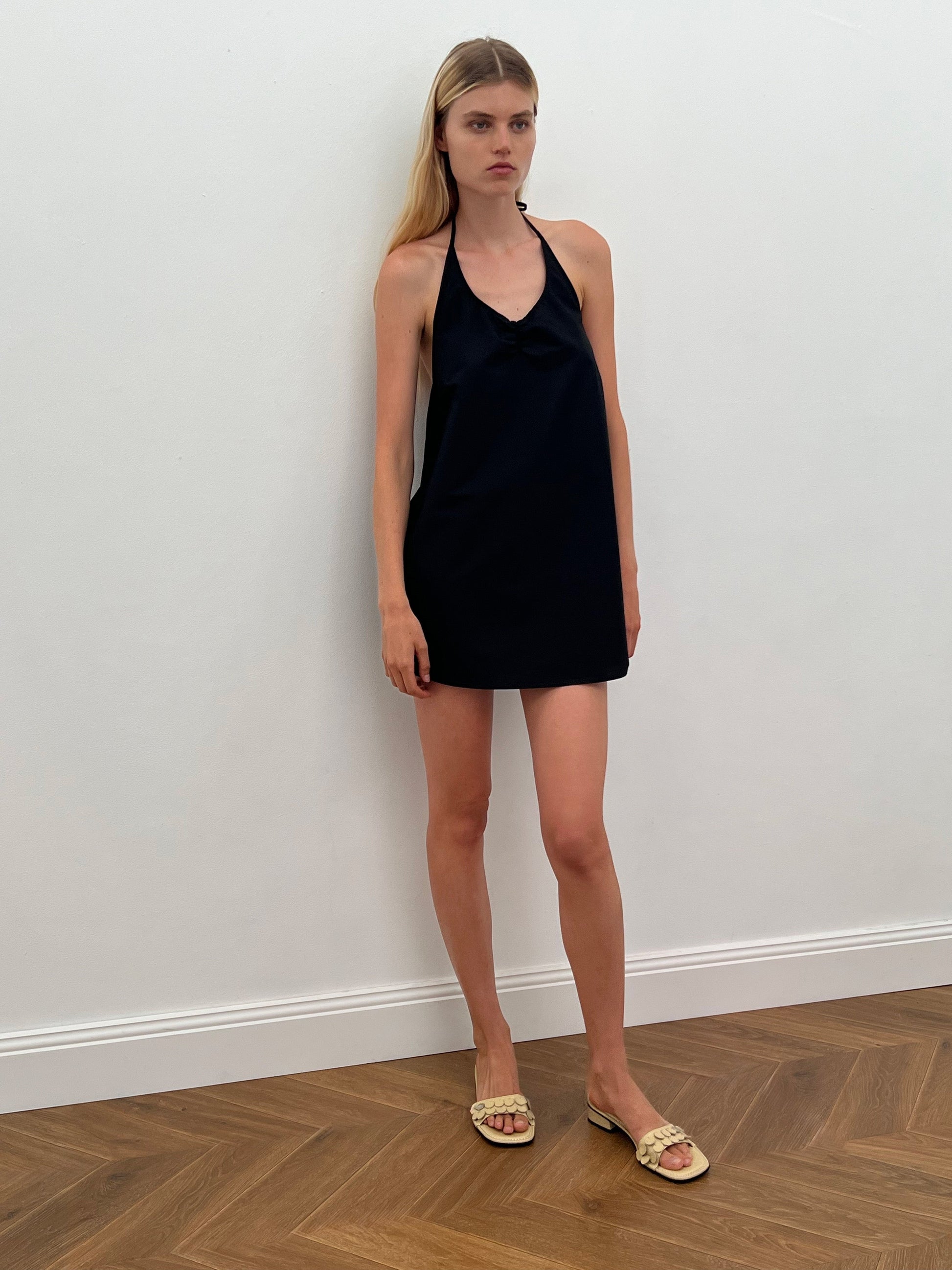A woman with long blonde hair stands against a white wall, wearing the Round Halter Dress - Black by 0 in organic cotton poplin and cream woven sandals, on a herringbone-patterned wooden floor.
