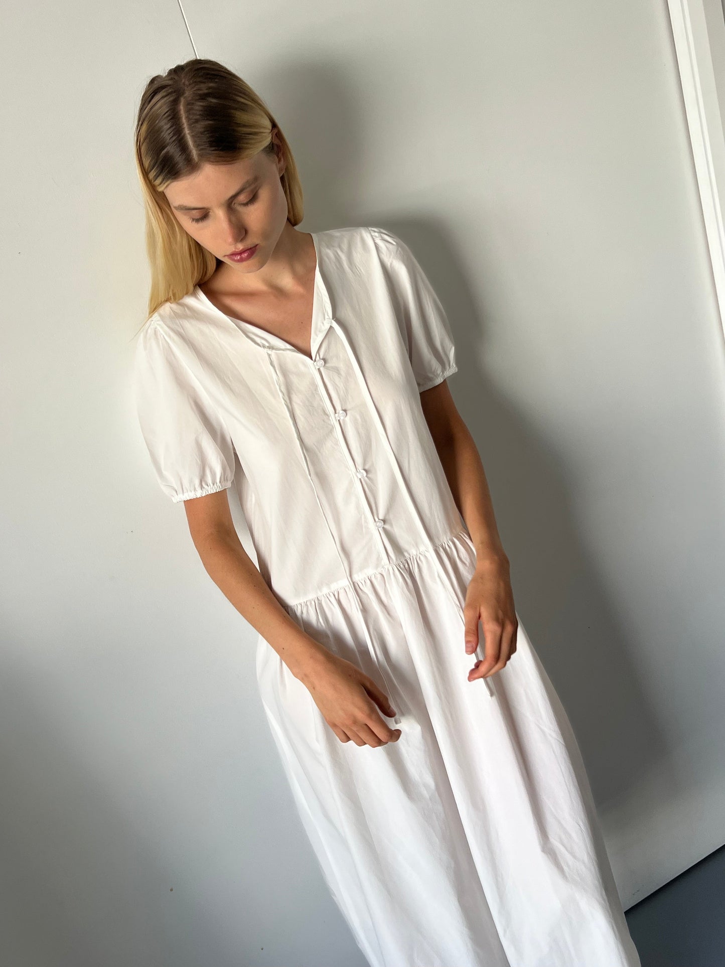 A woman with long blonde hair stands indoors against a light wall, wearing the Draw Hem Dress - White, a short-sleeved, buttoned dress, and looks down with a calm expression.