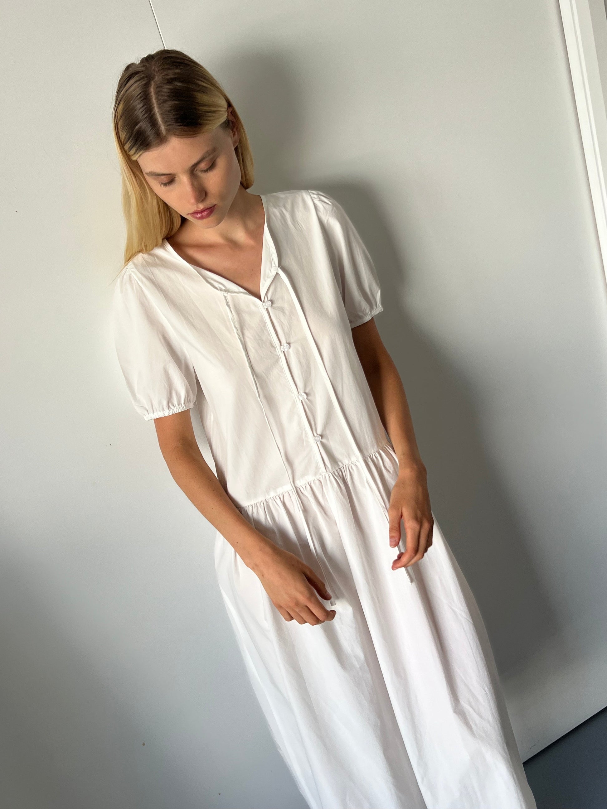 A woman with long blonde hair stands indoors against a light wall, wearing the Draw Hem Dress - White, a short-sleeved, buttoned dress, and looks down with a calm expression.